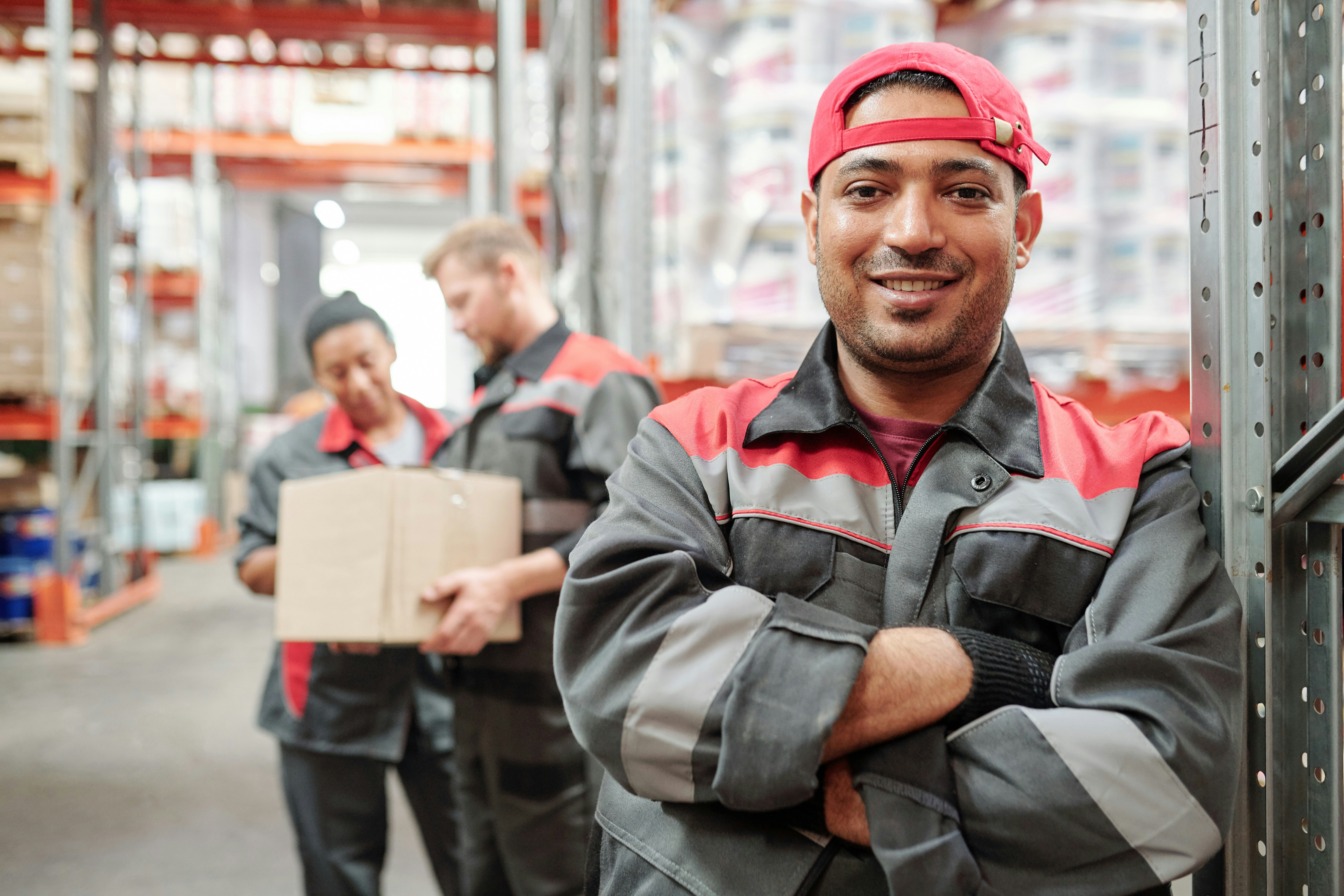 A man in grey and red workwear stands in a warehouse smiling at the camera; similarly dressed colleagues review paperwork in the background.