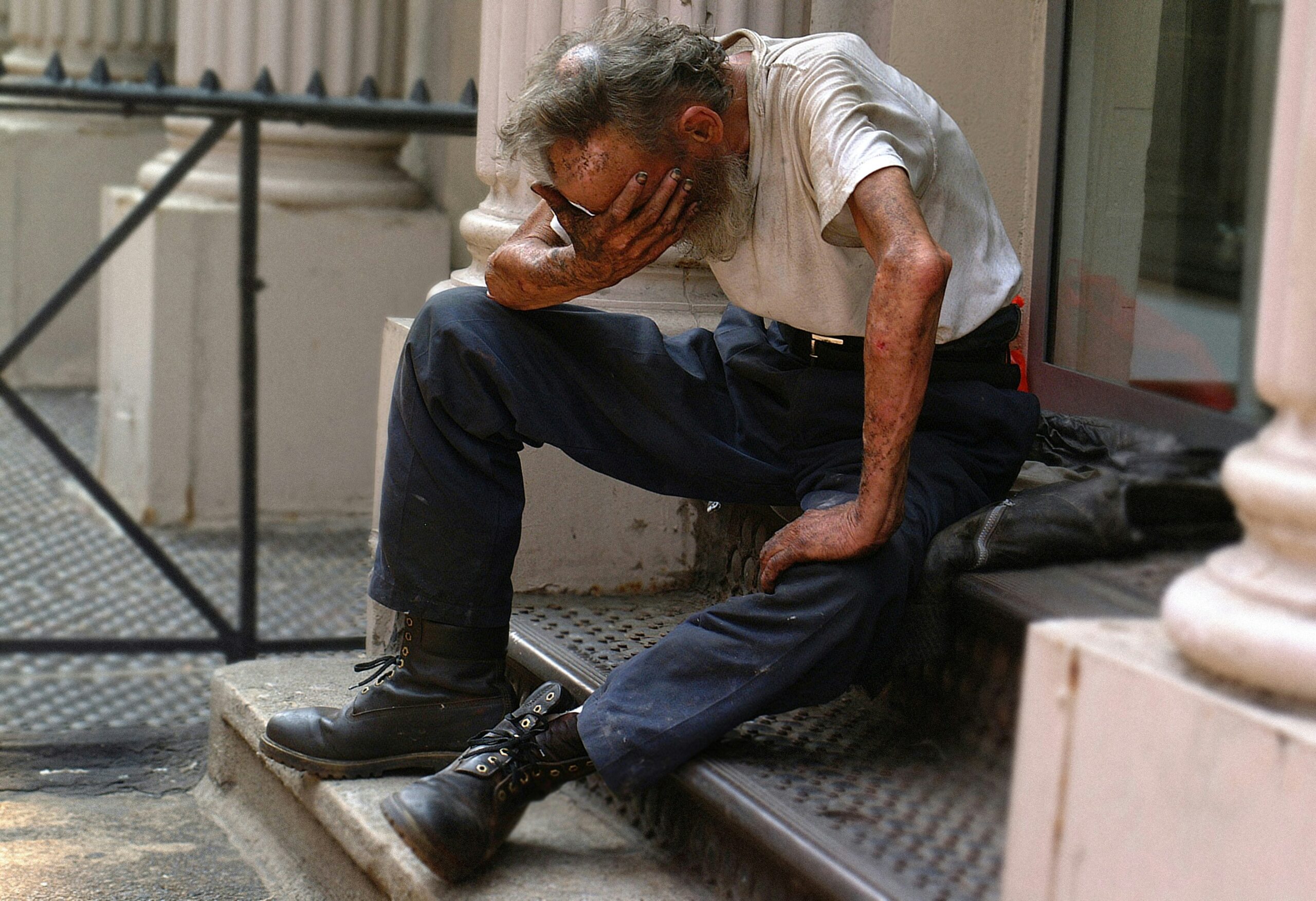 ben-hershey-8-fHqPCNy4c-unsplash A destitute man sits head in hand on steps outside of a columned doorway