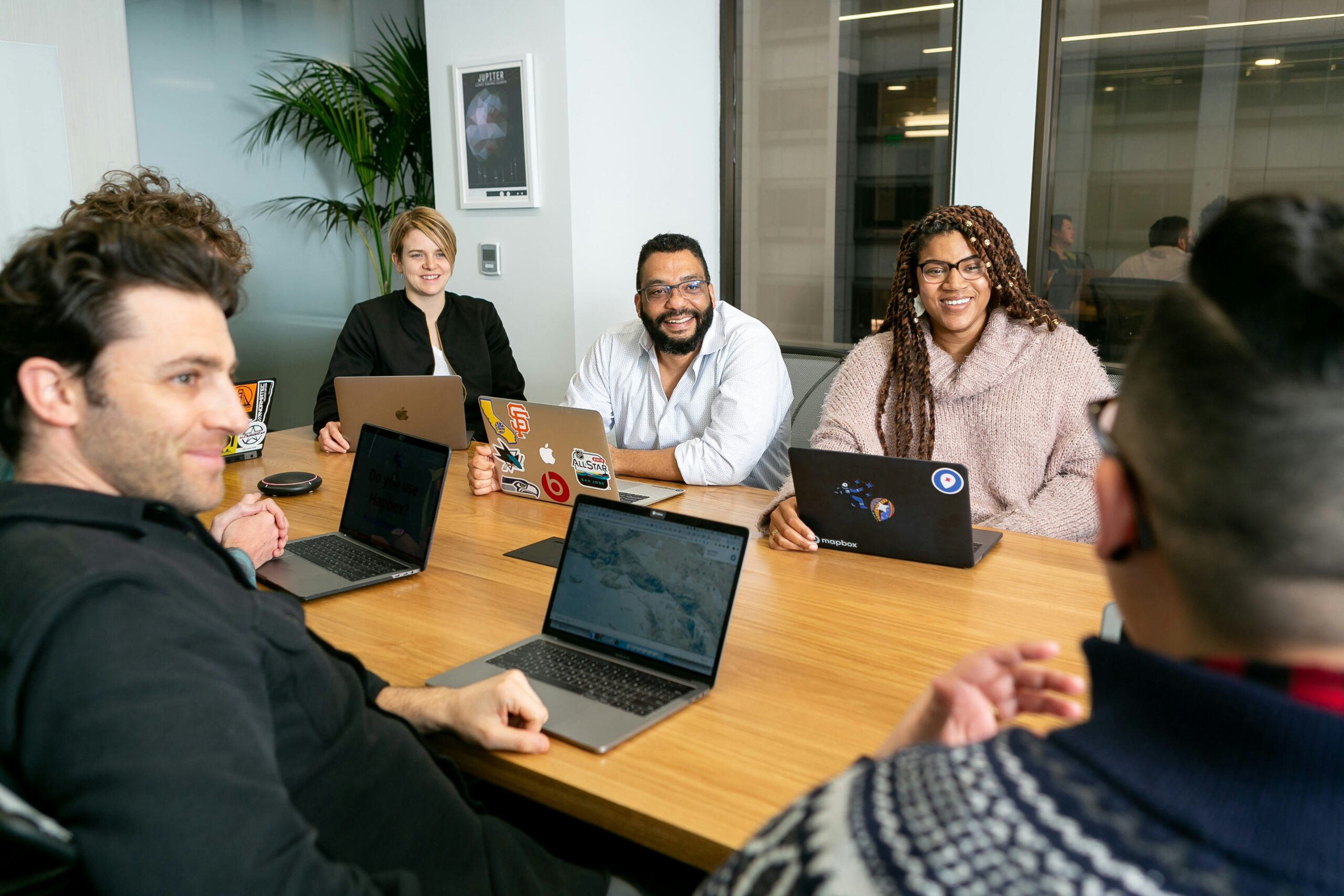 mapbox-ZT5v0puBjZI-unsplash A smiling group of office workers sits around a boardroom table with open laptops.
