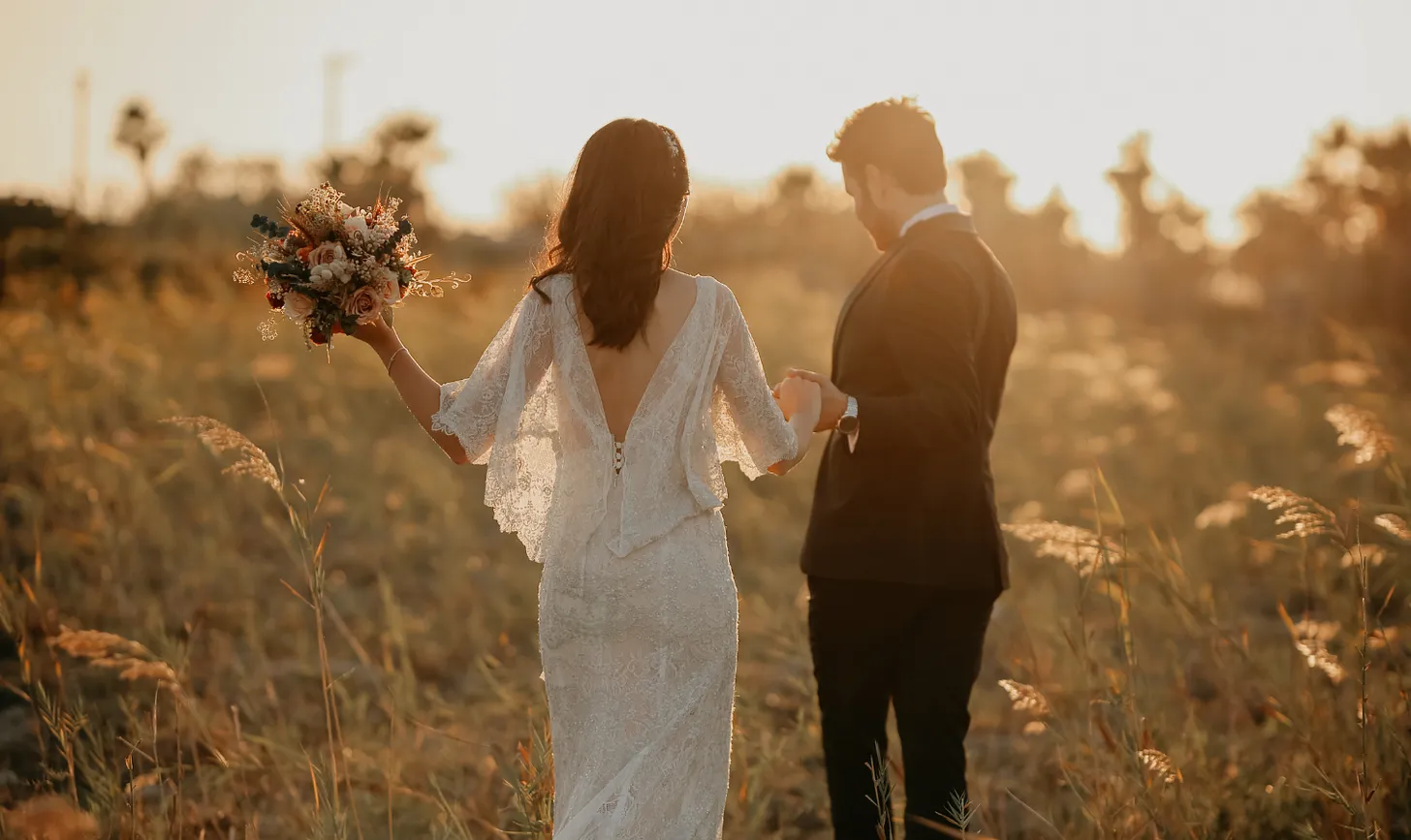 a bride and groom walk through a field in their wedding clothes at sunset