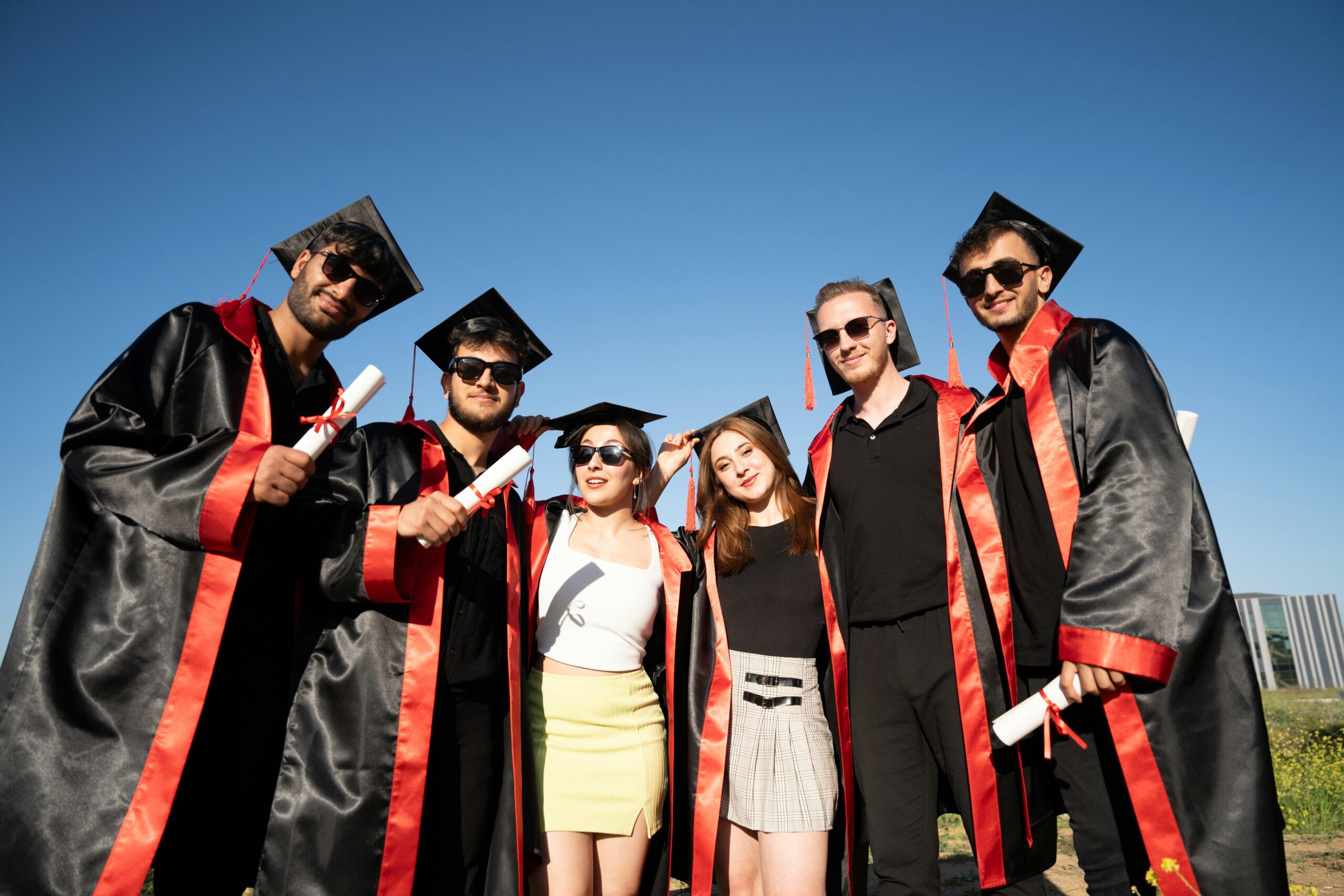 A group of recent graduates in black caps and gowns with red sashes smiles for the camera