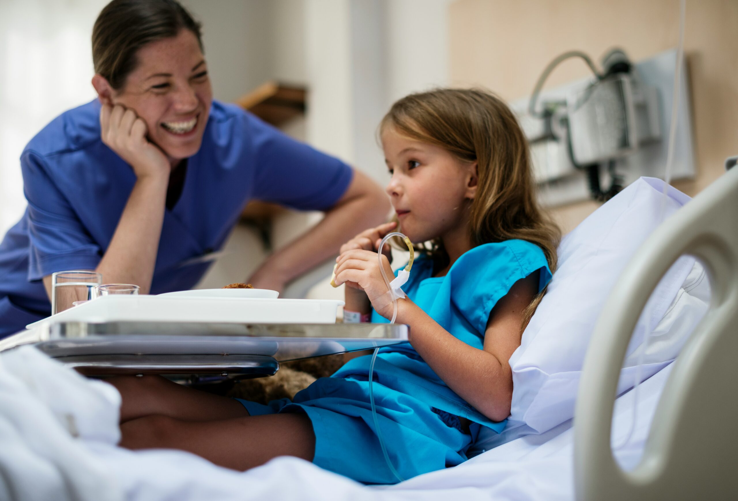 curated-lifestyle-X8mg6eMzGSA-unsplash A health care professional rests her head on her hand and smiles at a child eating in a hospital bed.