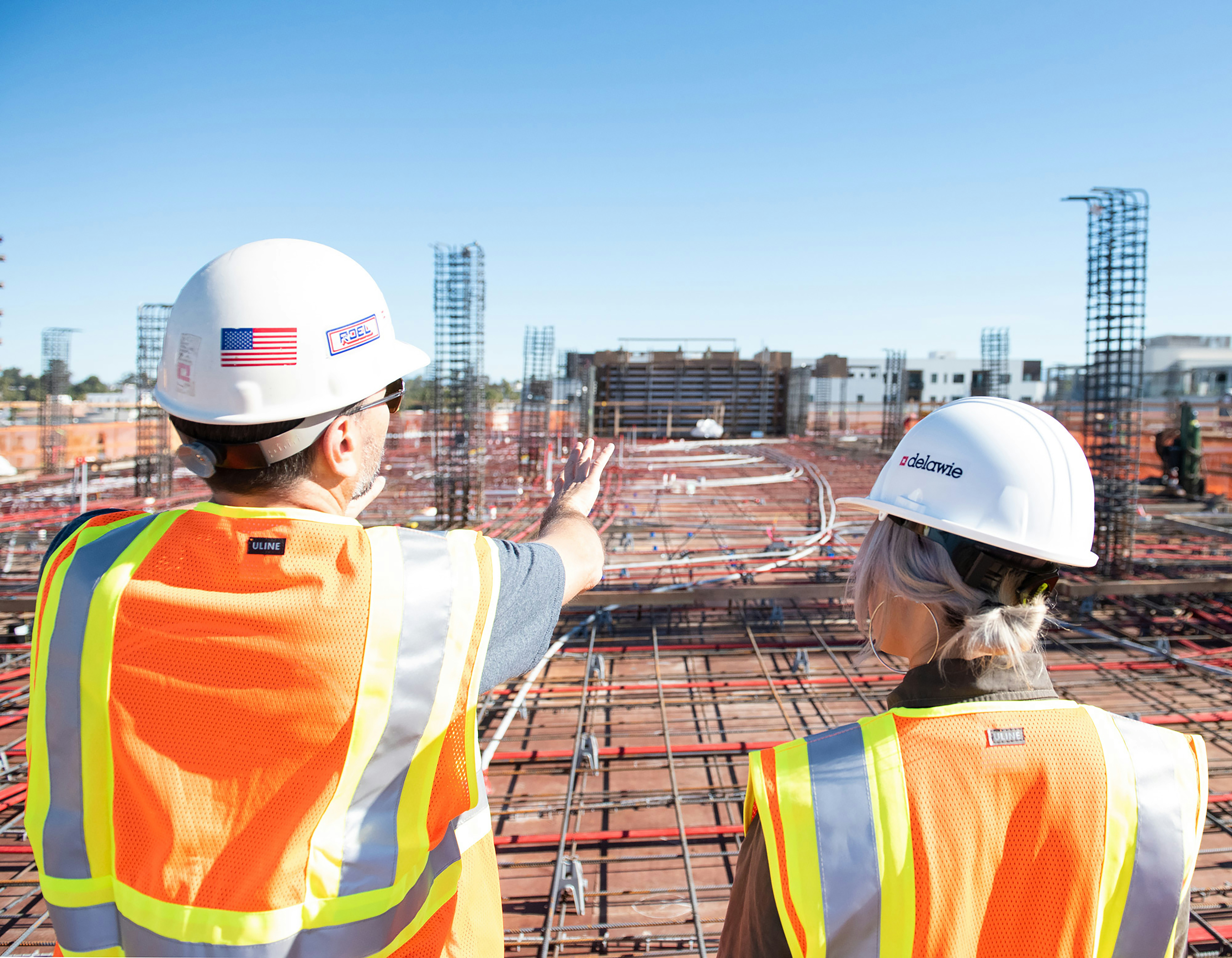 A man with an American-flag bedecked hard hat and reflective safety vest points across a large work project saying something to a smiliarly adorned woman
