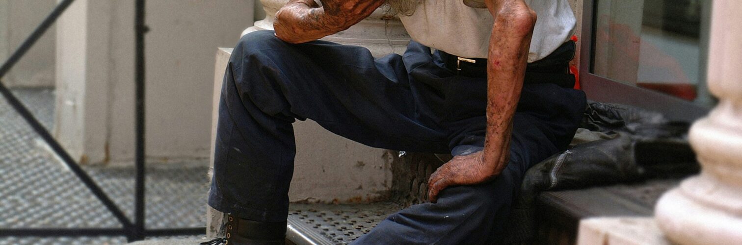 A destitute man sits head in hand on steps outside of a columned doorway