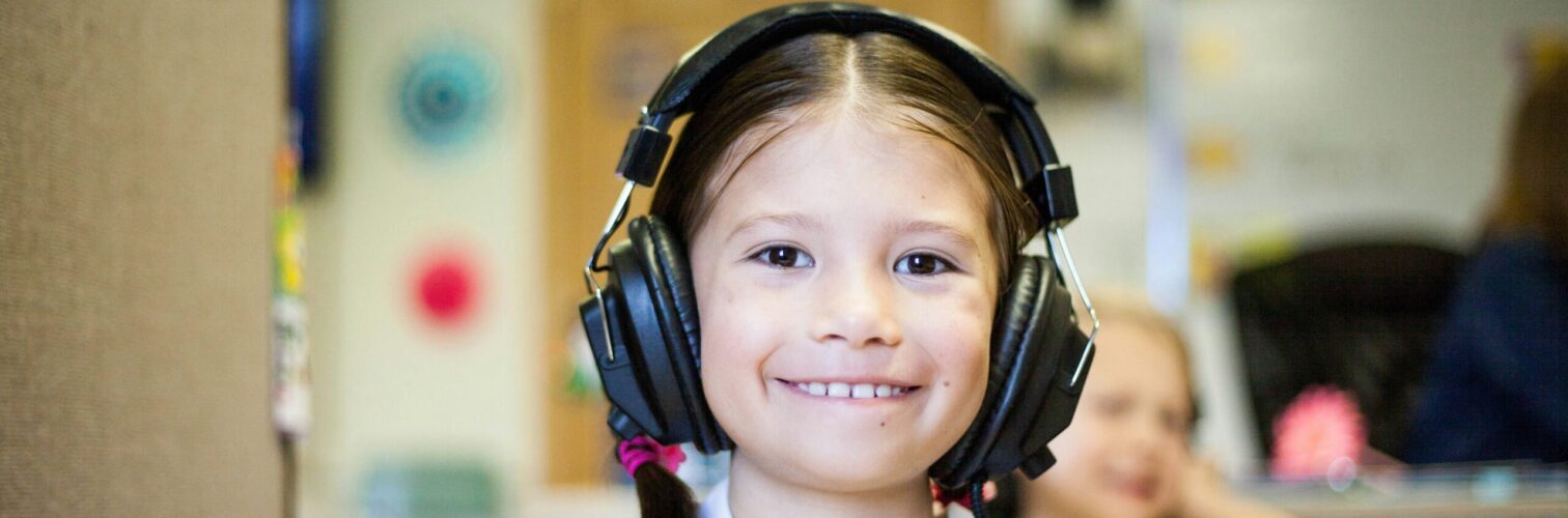 A young girl wearing headphones smiles in a classroom.