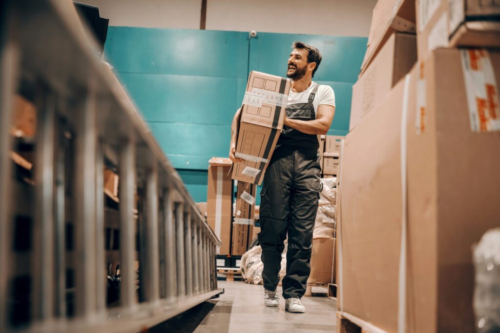 A man with a beard in denim coveralls and a white t-shirt carries a large cardboard box through a warehouse