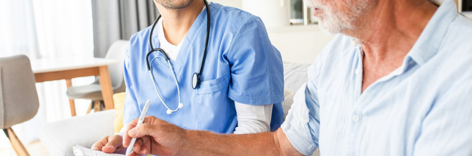 An older patient sitting on his bed signs paperwork for his smiling health care provider