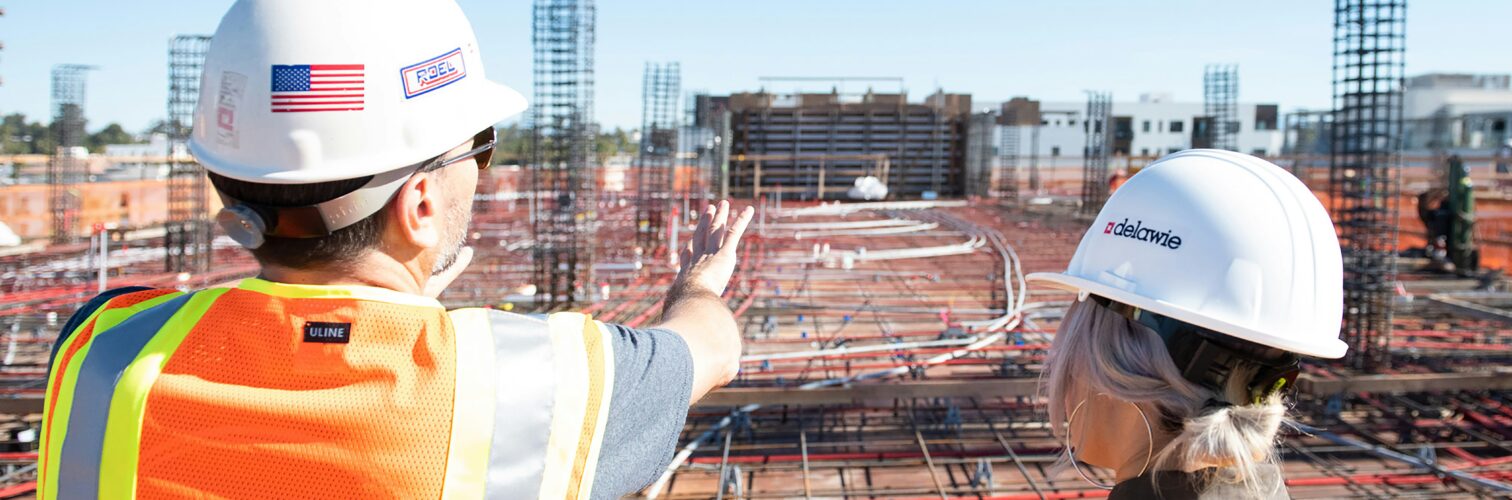 A man with an American-flag bedecked hard hat and reflective safety vest points across a large work project saying something to a smiliarly adorned woman