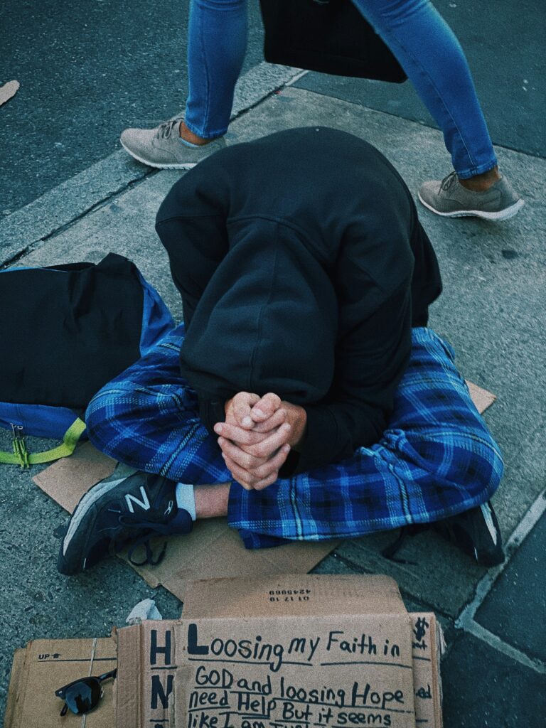 A homelss man in a hoodie sits crosslegged with his hands clasped in front of him in behind a sign asking for help