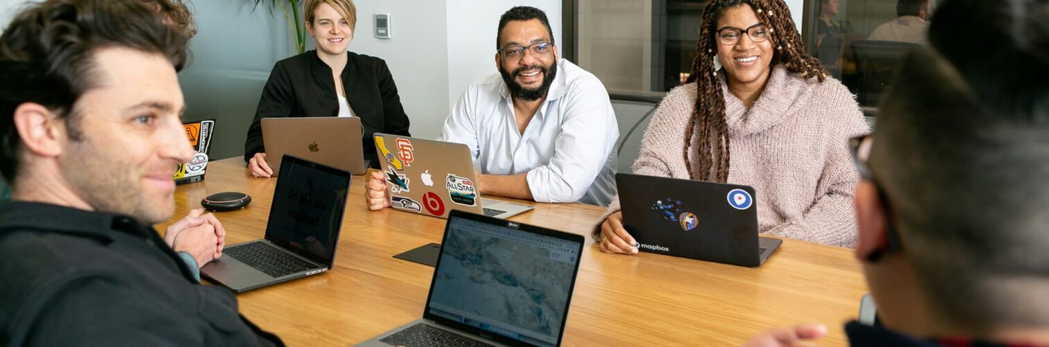 A smiling group of office workers sits around a boardroom table with open laptops.