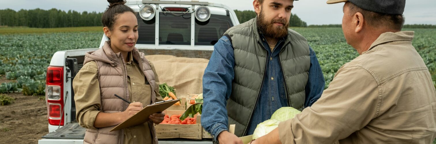 A man hands another man a box of produce in front of a pickup truck with the tailgate down. They are in a farm field and a woman with her hair in a bun looks on. Photo courtesy of Getty Images via Unsplash.