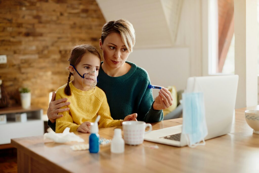 A mother holds a thermometer in one hand and her daughter wearing an oxygen mask on her lap in the other while looking at a laptop, probably in a digitial doctor visit