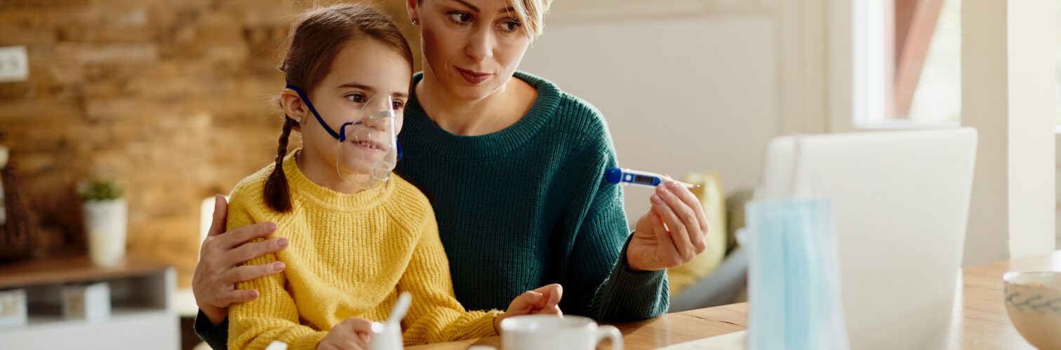 A mother holds a thermometer in one hand and her daughter wearing an oxygen mask on her lap in the other while looking at a laptop, probably in a digitial doctor visit