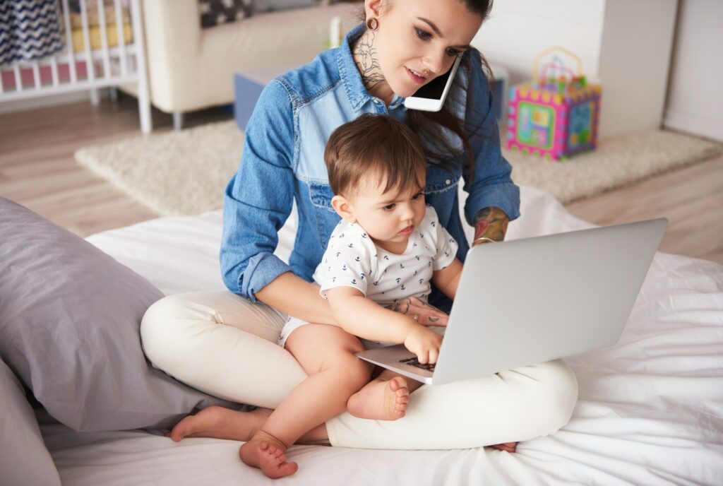 A mom with a neck tattoo sits cross legged with her toddler and laptop on her lap while talking on the phone.