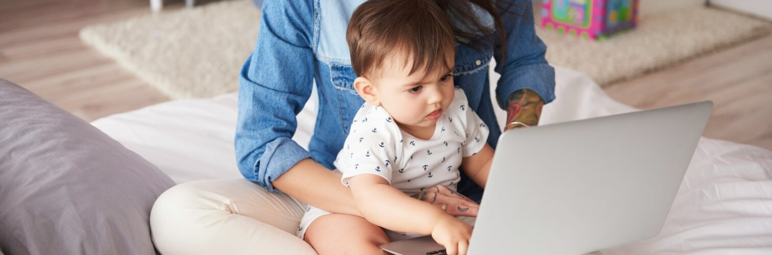 A mom with a neck tattoo sits cross legged with her toddler and laptop on her lap while talking on the phone.