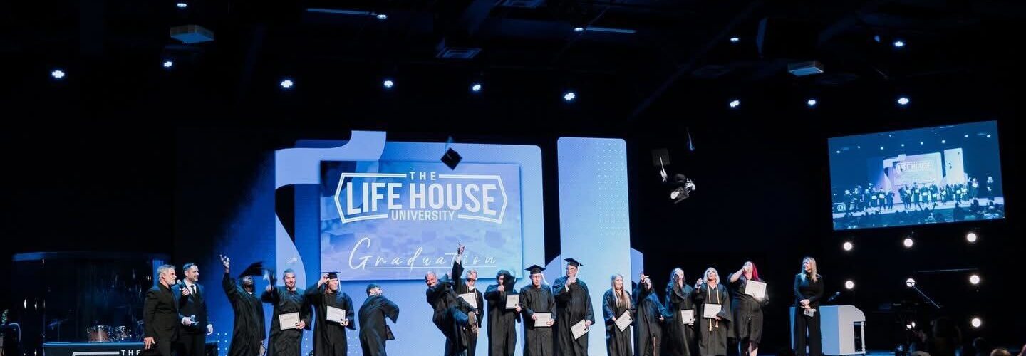 Ecstatic graduates throw their caps into the air on stage in front of of a large sign that reads “The Life House Graduation"